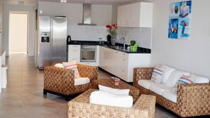 Une cuisine moderne avec des meubles blancs, un réfrigérateur en acier inoxydable et un plan de travail noir. Le salon est aménagé avec des canapés en osier dotés de coussins blancs et une petite table.