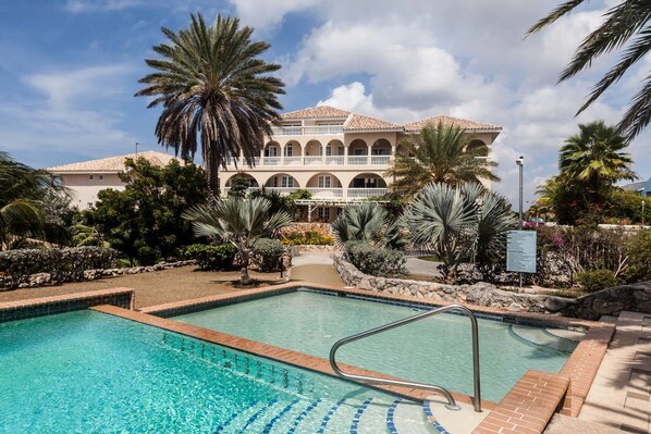 A pool area with a clear blue pool, surrounded by palm trees and a building with arched windows and a tiled roof.