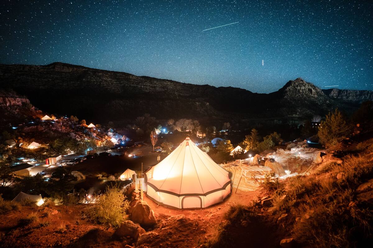 A large, illuminated yurt surrounded by a campsite with tents and a starry night sky.