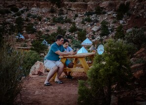 Duas pessoas sentadas numa mesa de piquenique de madeira ao ar livre, rodeadas por terreno rochoso e vegetação escassa.