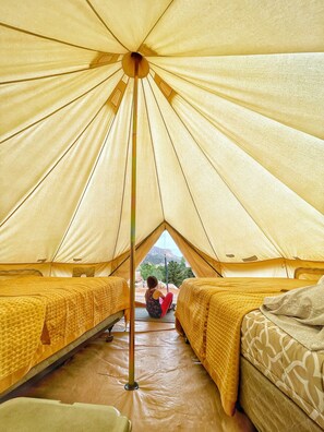 Uma tenda com um mastro central, duas camas com roupa de cama amarela e vista para uma paisagem montanhosa pela entrada.