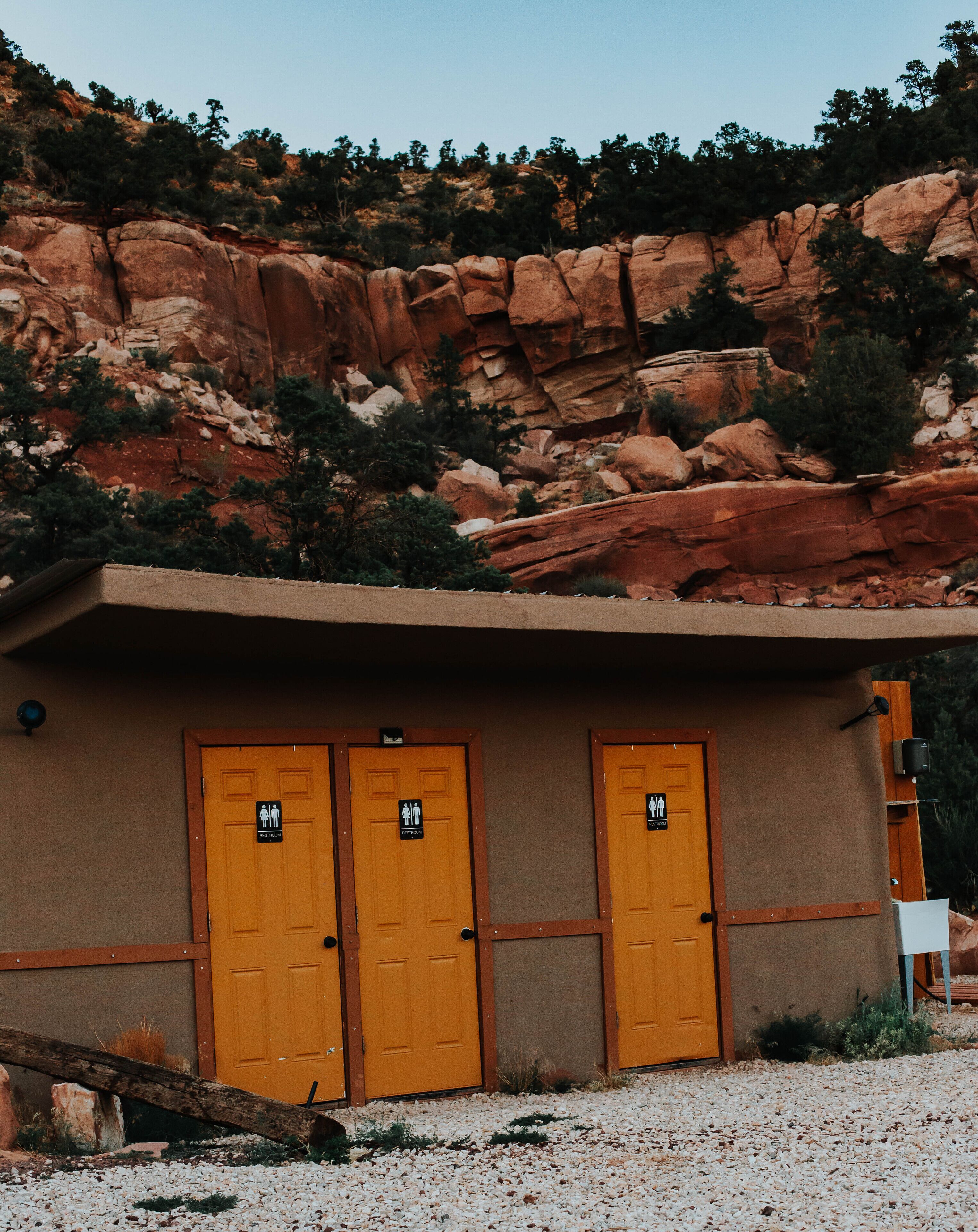 A building with three wooden doors, each marked with a restroom sign. The building is set against a backdrop of red rock formations and sparse vegetation.