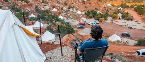 En person sidder ved et bord uden for et telt og kigger ud over en campingplads med flere telte og køretøjer.