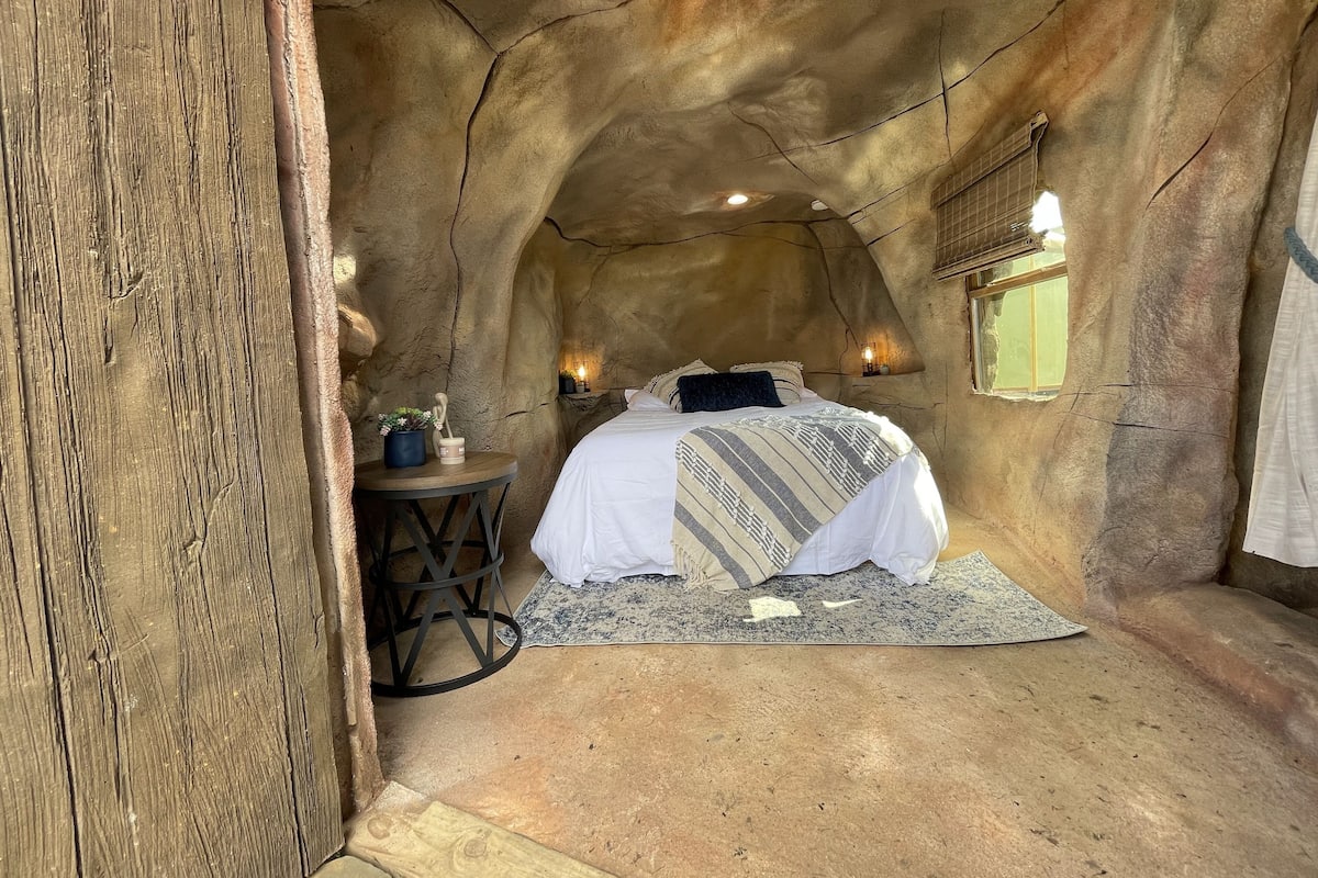 A cozy, cave-like bedroom with a bed, a small table with a plant, and a wall-mounted light fixture.