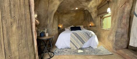 A cozy, cave-like bedroom with a bed, a small table with a plant, and a wall-mounted light fixture.