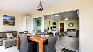 A dining area with a wooden table and wicker chairs, a sofa, and a view through a glass door to another room. - Ocean Resort Villa Condor (Willemstad)