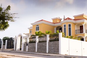 A large, two-story house with a yellow facade, white trim, and a red-tiled roof. It features a gated entrance, a stone wall, and a balcony with a railing. - Villa with Spectacular Sea View (Jan Thiel)