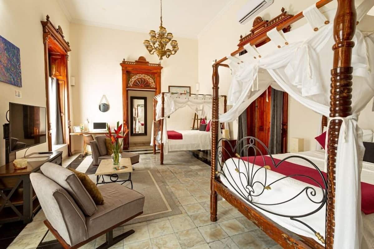 A spacious bedroom with a four-poster bed, a sitting area with a sofa and armchair, a television, and a desk.