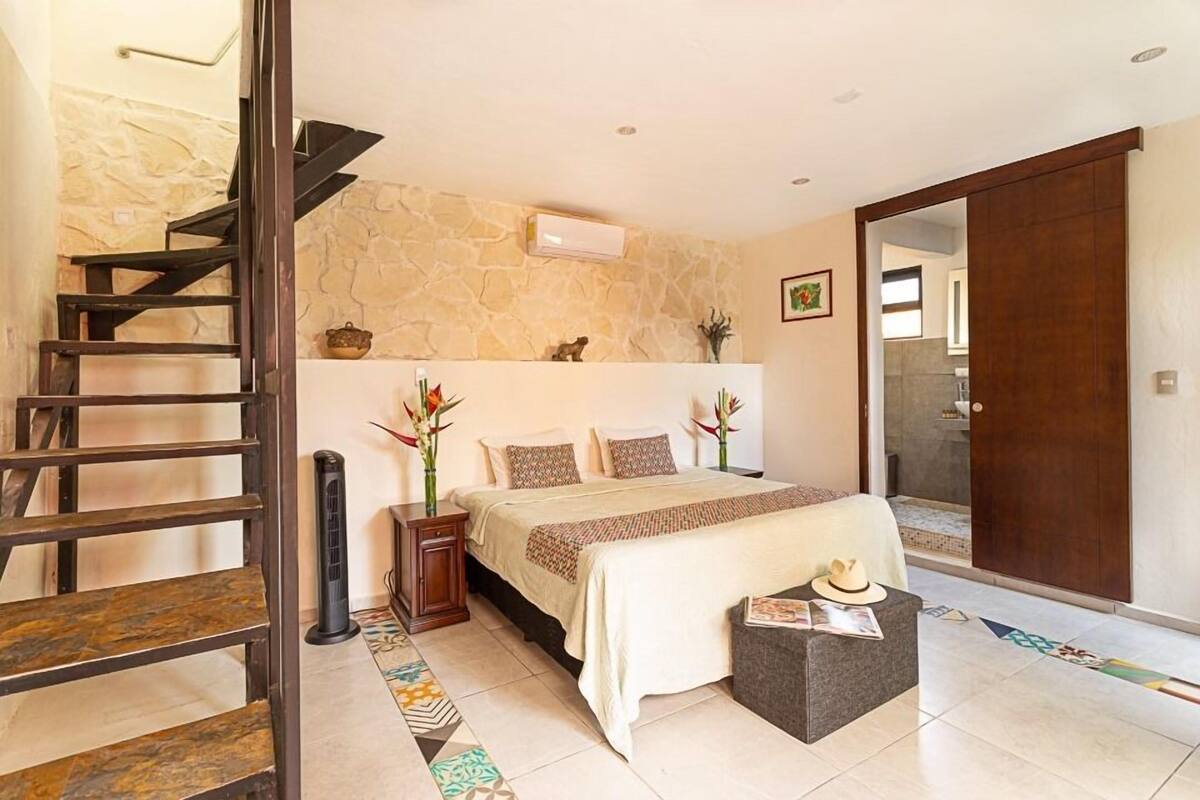 A bedroom with a bed, a nightstand, a bedside table, a chair, a wooden staircase, and a door.
