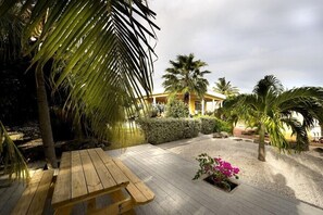 A wooden deck with a bench, surrounded by palm trees and a hedge, leading to a building with a yellow facade.