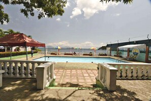 Zona de piscina con una toldo rojo, sombrillas y vista al mar.