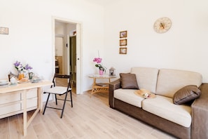 Un salon avec un canapé beige, une table à manger en bois avec des chaises, une petite table d’appoint sur laquelle se trouve un vase rose, et une horloge murale.
