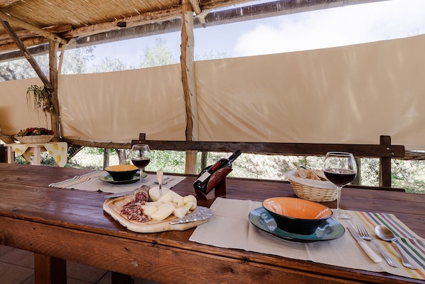 Une table en bois, dressée avec des assiettes, des bols, des couverts et une bouteille de vin, placée sous une zone abritée d’où l’on peut admirer la vue sur les arbres.