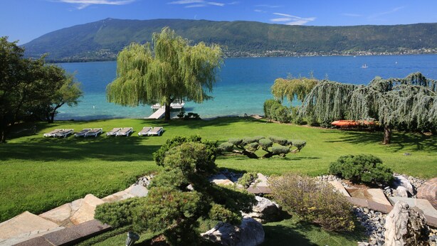 Un jardín tranquilo junto al lago con un árbol, sillas de descanso y una vista despejada del lago y las montañas.