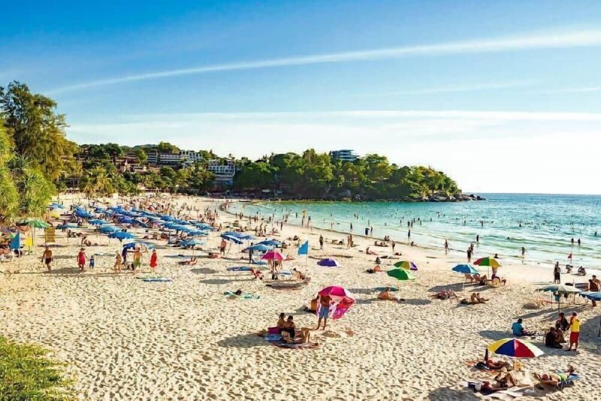 A crowded beach with people sunbathing and swimming, umbrellas, and buildings in the background.