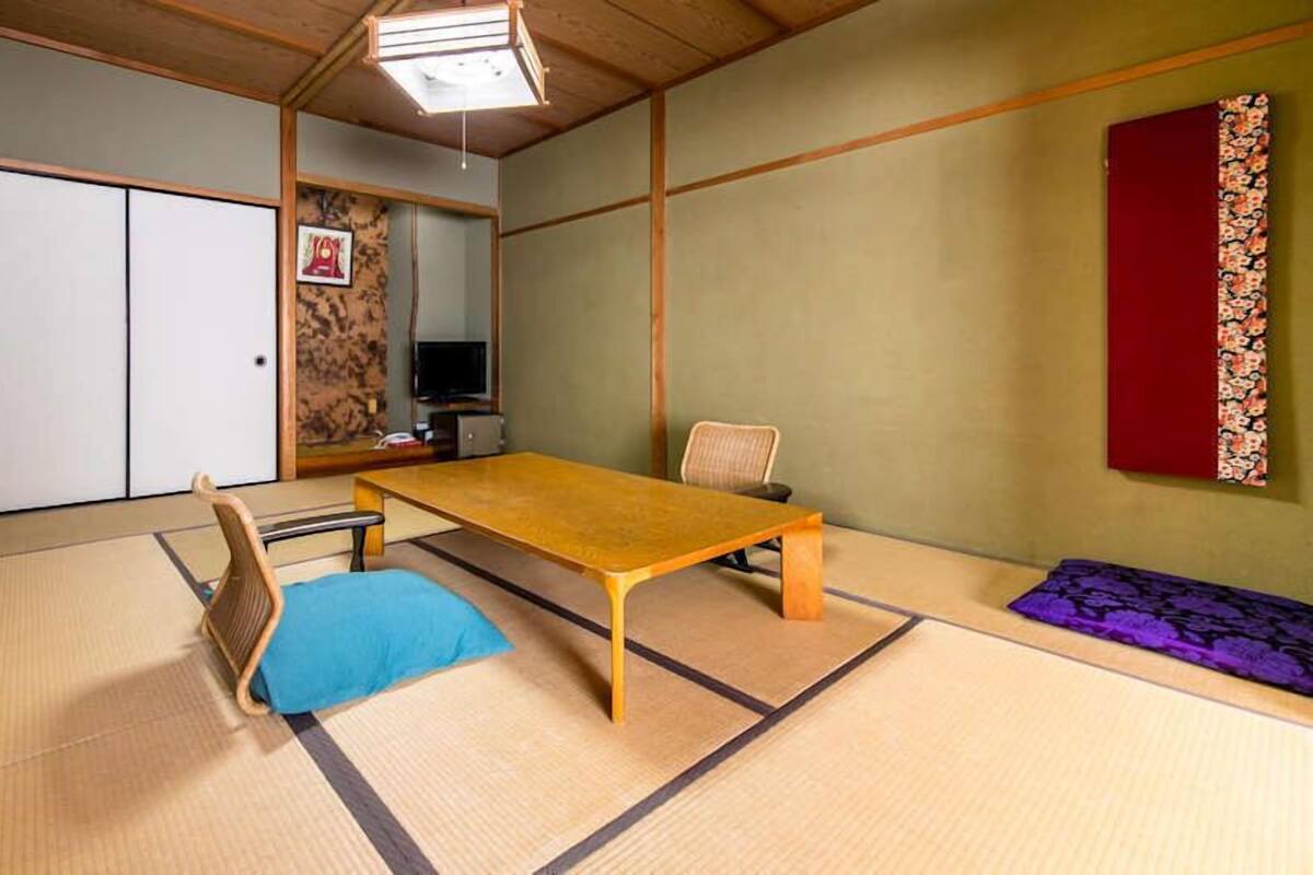 A tatami mat room with a low table, a chair, a television, and a red curtain.