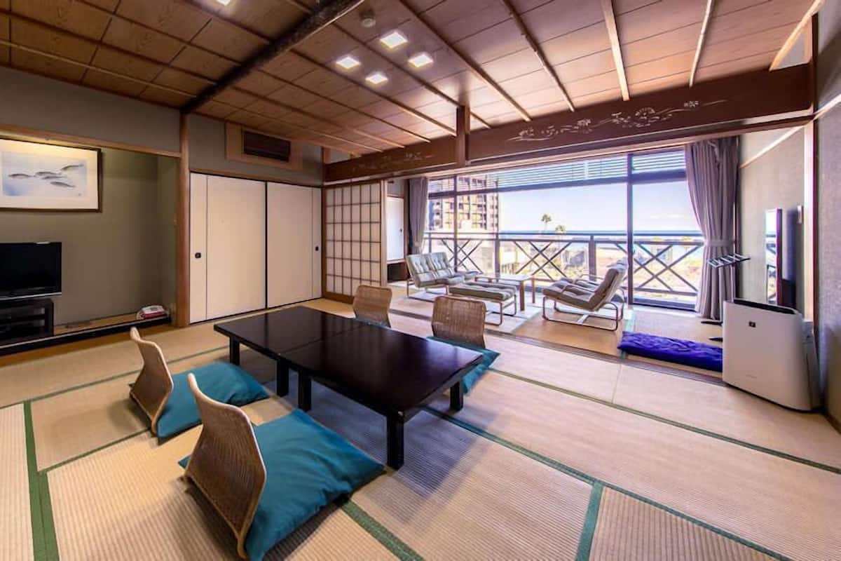 A room with tatami mats, a low table, chairs, a television, and a balcony with sun loungers.