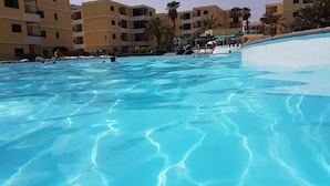 Ein klares Schwimmbecken mit sichtbaren Wellen, umgeben von gelben Gebäuden mit Balkonen und Palmen.