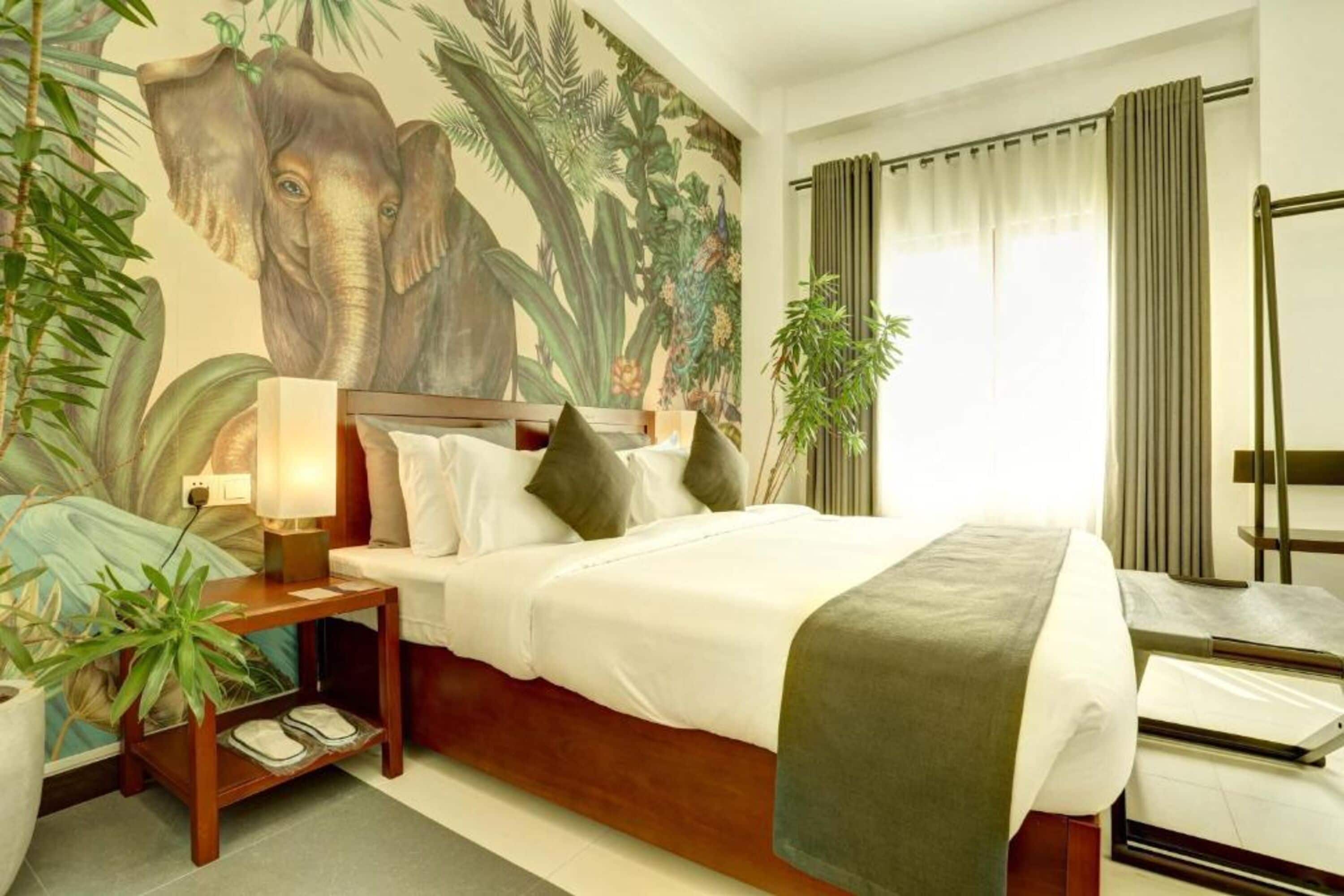Habitación de hotel con un gran mural de elefante, una cama con sábanas blancas, una mesita de noche con una lámpara y una planta en maceta.
