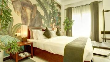 Habitación de hotel con un gran mural de elefante, una cama con sábanas blancas, una mesita de noche con una lámpara y una planta en maceta.