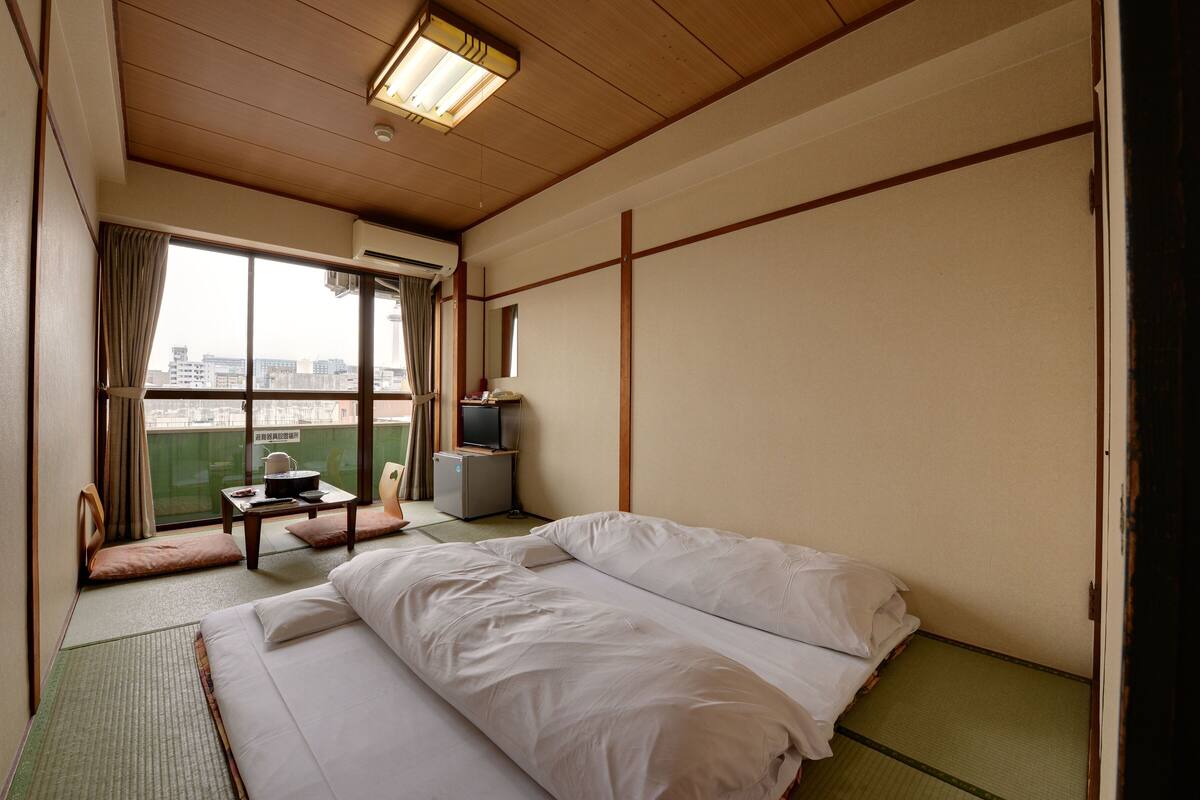 Habitación de hotel con cama, una mesita con silla, televisión y vistas a la ciudad a través de un ventanal grande.