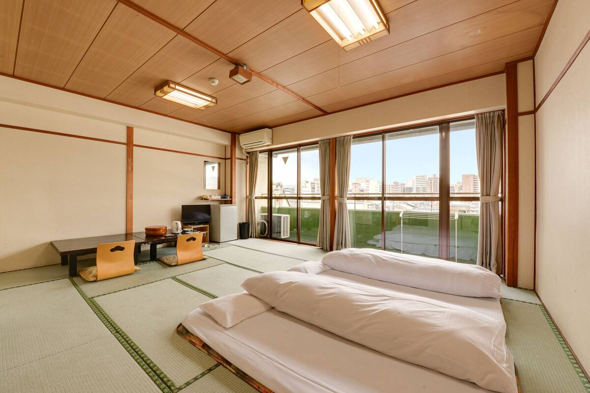 Habitación de hotel con suelo de tatami, una cama, un escritorio con silla, un televisor y un ventanal con vistas a la ciudad.