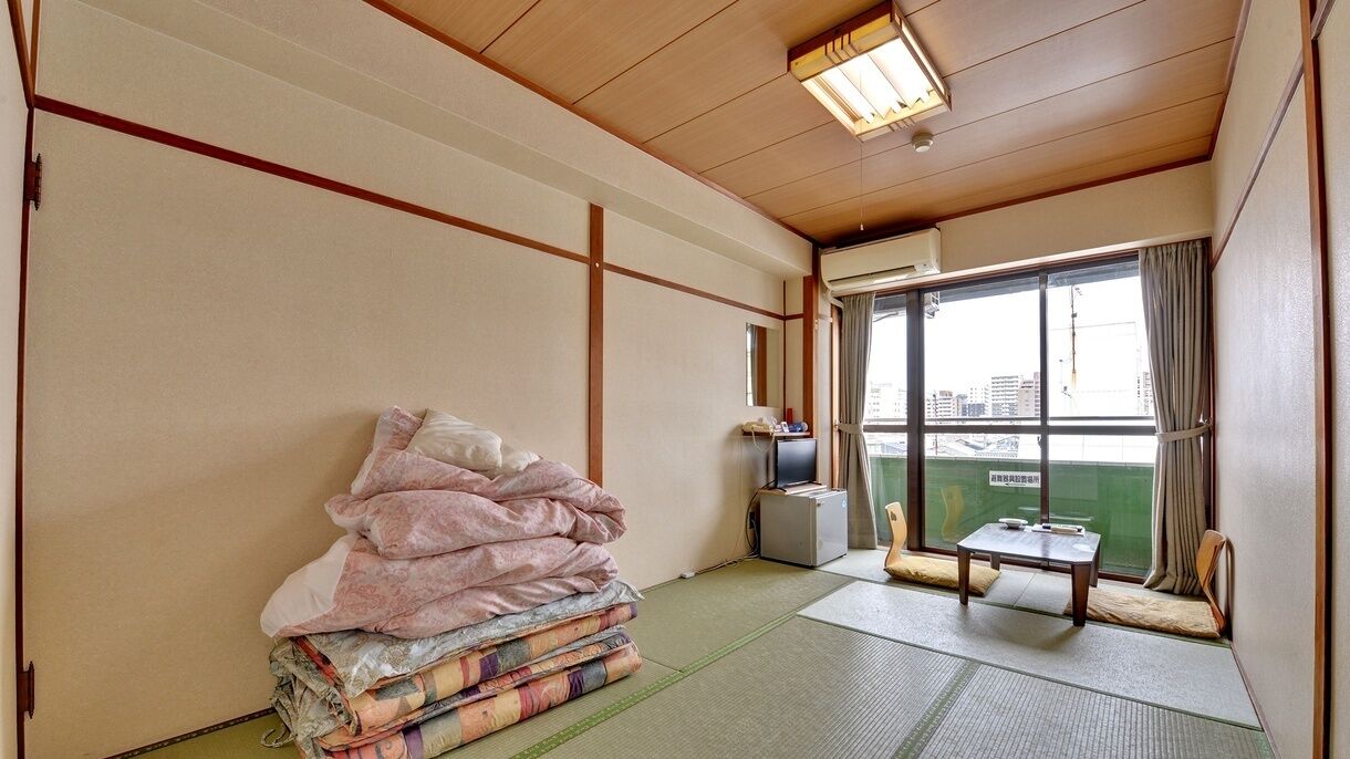 A room with tatami flooring, a futon bed, a television, and a view of a cityscape through a large window.