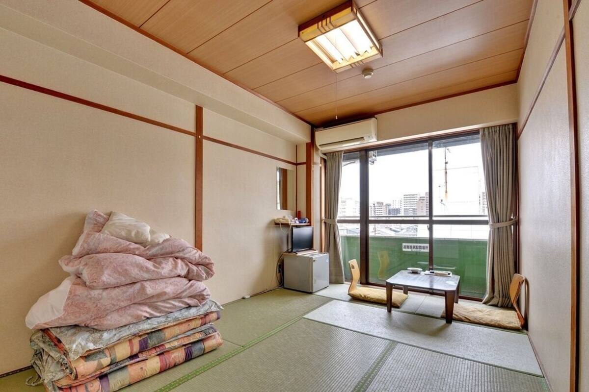 Habitación con suelo de tatami, futón, televisión y vistas a un paisaje urbano a través de un ventanal grande.