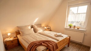 A bedroom with a sloped ceiling, a wooden bed, bedside tables with lamps, a window with plants, and a radiator.