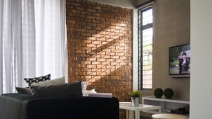 Ein modernes Wohnzimmer mit einer schwarzen Couch, einem kleinen weißen Tisch, einem an der Wand befestigten Fernseher und einer Ziegelwand als Blickfang.
