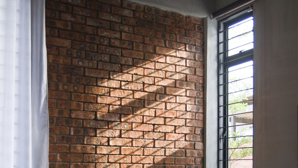 Ein Zimmer mit Backsteinwand, einem weißen Stuhl, einem kleinen runden Tisch und einem Fenster mit Gitterstäben.