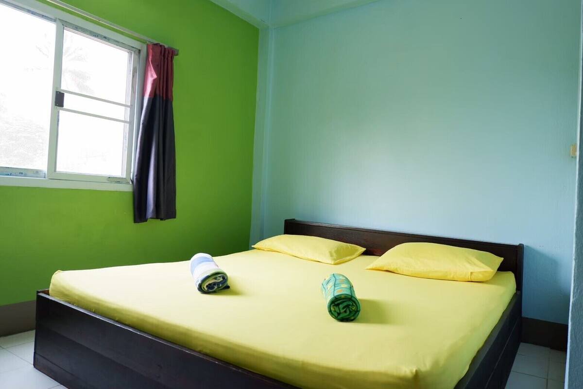 Una cama individual con ropa de cama amarilla, dos almohadas y dos toallas enrolladas en una habitación con paredes verdes y una ventana con cortinas.