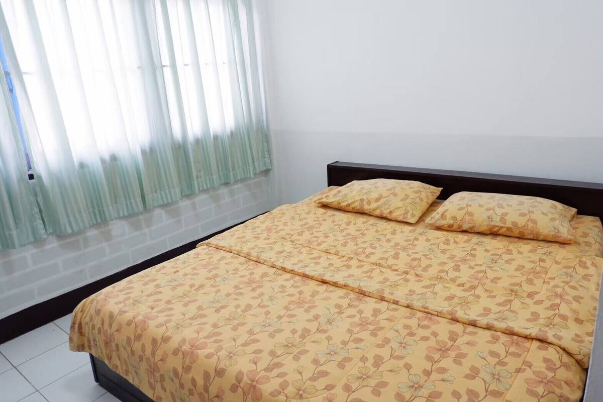Una cama con ropa de cama floral, una ventana con cortinas traslúcidas y una pared blanca lisa.