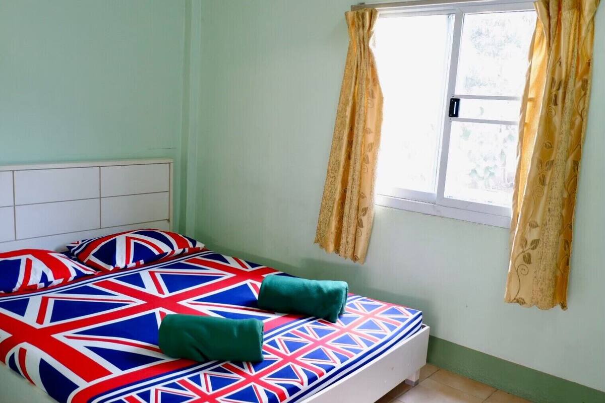 Una cama individual con una colcha y almohadas estampadas con la bandera del Reino Unido, un plaid verde y una ventana con cortinas amarillas.