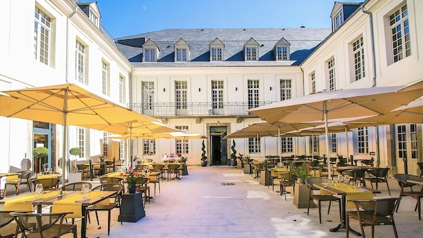 An outdoor cafe with tables and chairs, surrounded by a building with large windows and a balcony. - Charmant T2, hypercentre, calme, terrasse, parking, commerces (Bagnères-De-Luchon)