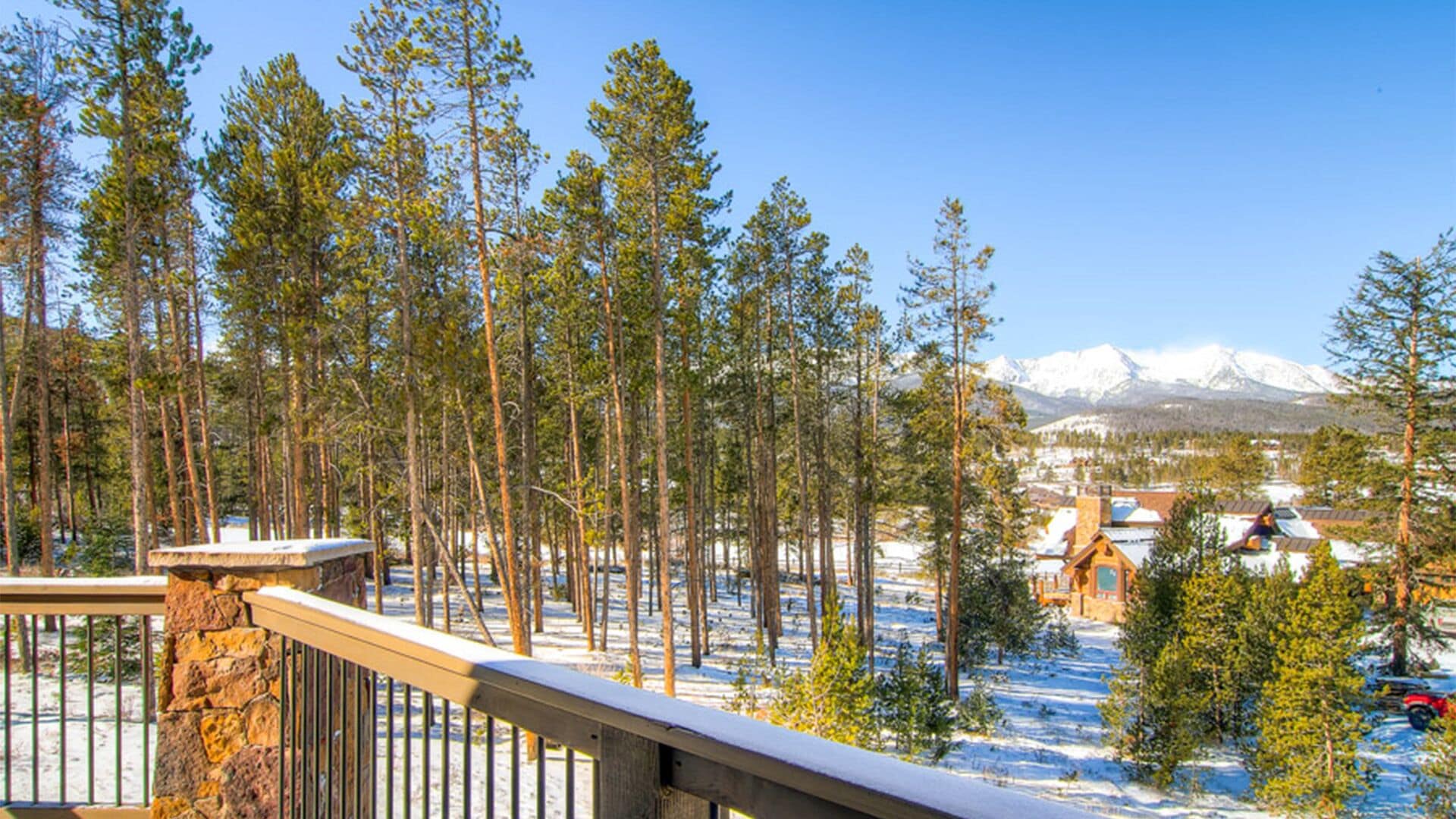A snowy landscape with tall pine trees, a stone railing, and a view of a small town with buildings and snow-covered mountains in the background.