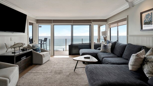Een ruime woonkamer met een grote flatscreen-tv, een grijze bank, een bijzettafeltje en uitzicht op de oceaan door middel van grote schuifdeuren.