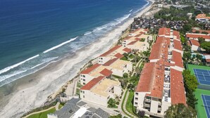Luchtbeeld van een kustgebied met woningen direct aan het strand, een tennisbaan en een helder uitzicht op de oceaan.