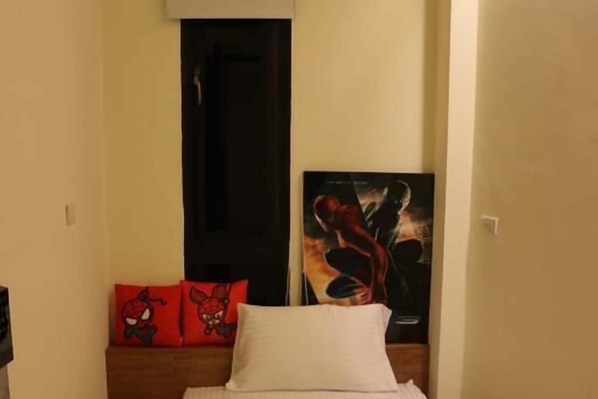 Una cama individual con colchón blanco y una manta gris doblada. Hay un almohadón rojo con un personaje de dibujos animados en el cabecero. Se ve una puerta negra al fondo.