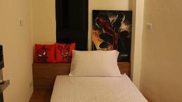 A single bed with a white mattress and a folded gray blanket. There is a red pillow with a cartoon character on the headboard. A black door is visible in the background.