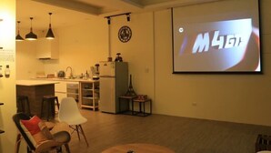 Una sala de estar moderna con una zona de cocina, una pantalla de proyector mostrando "M4GT" y un reloj en la pared.