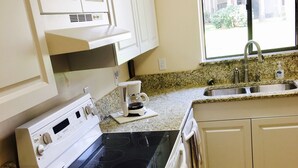 A kitchen with white cabinets, a stove, a coffee maker, and a window with a view of trees.