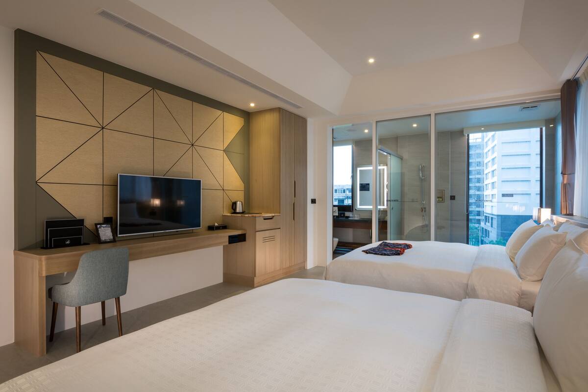 Une chambre d’hôtel moderne dotée d’un grand lit, d’un bureau avec un téléviseur à écran plat, d’un coin salon et d’une porte vitrée donnant sur un balcon avec vue sur la ville.