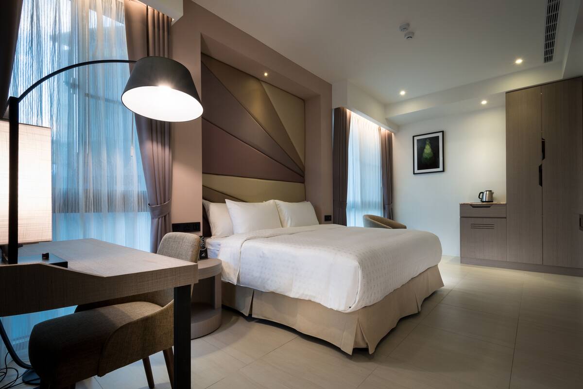Une chambre d’hôtel moderne équipée d’un lit, d’un bureau, d’une chaise, d’une télévision et d’une petite kitchenette.