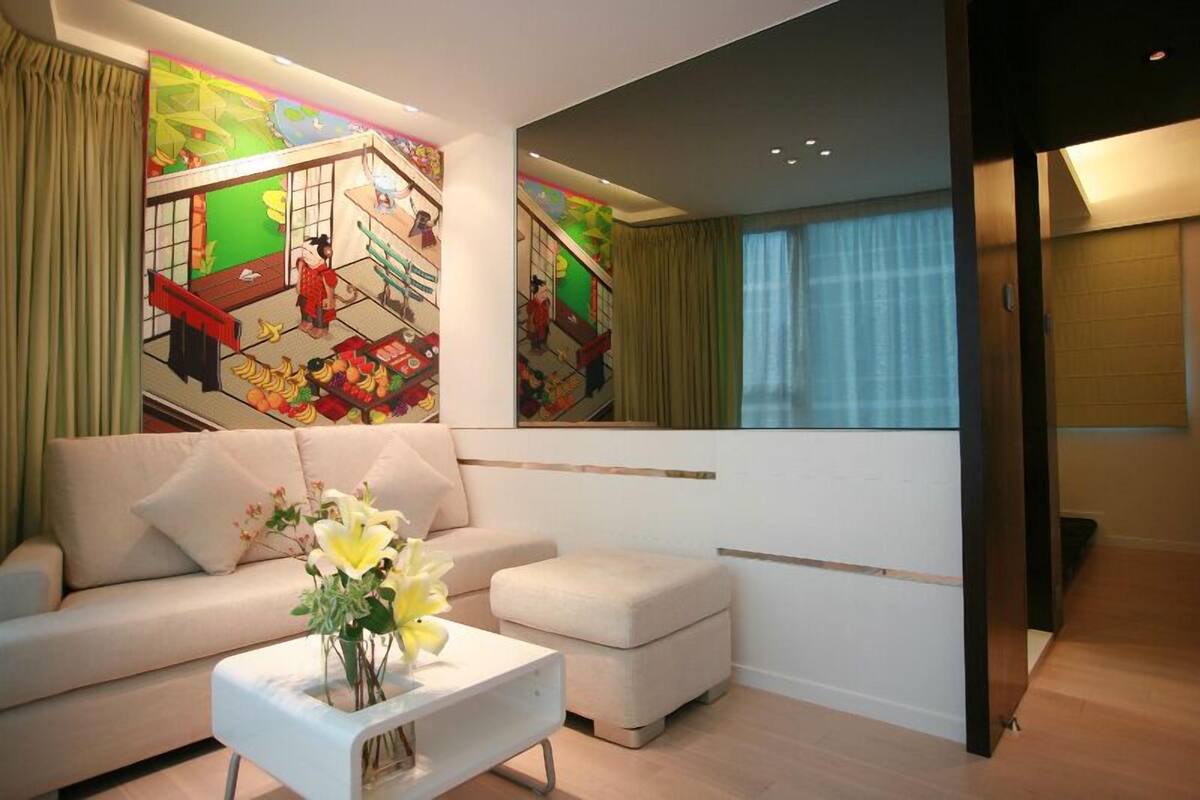 Een moderne woonkamer met een bank, een poef en een salontafel met bloemen. Een kleurrijke muurschildering en een groot raam met gordijnen zijn zichtbaar.