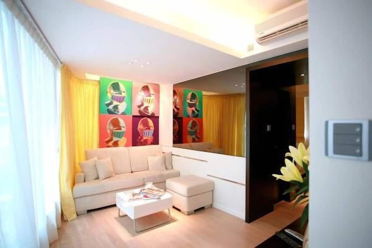 Een moderne woonkamer met een bank, een poef en een salontafel. Op de muur hangen kleurrijke abstracte schilderijen en er is een grote spiegel.