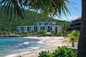 A beachfront hotel with multiple floors, a sandy beach, and a backdrop of lush green hills. - Romantic Oceanview Escape -- Private Balcony Views -- Close to Sapphire Beach (Sapphire Village)