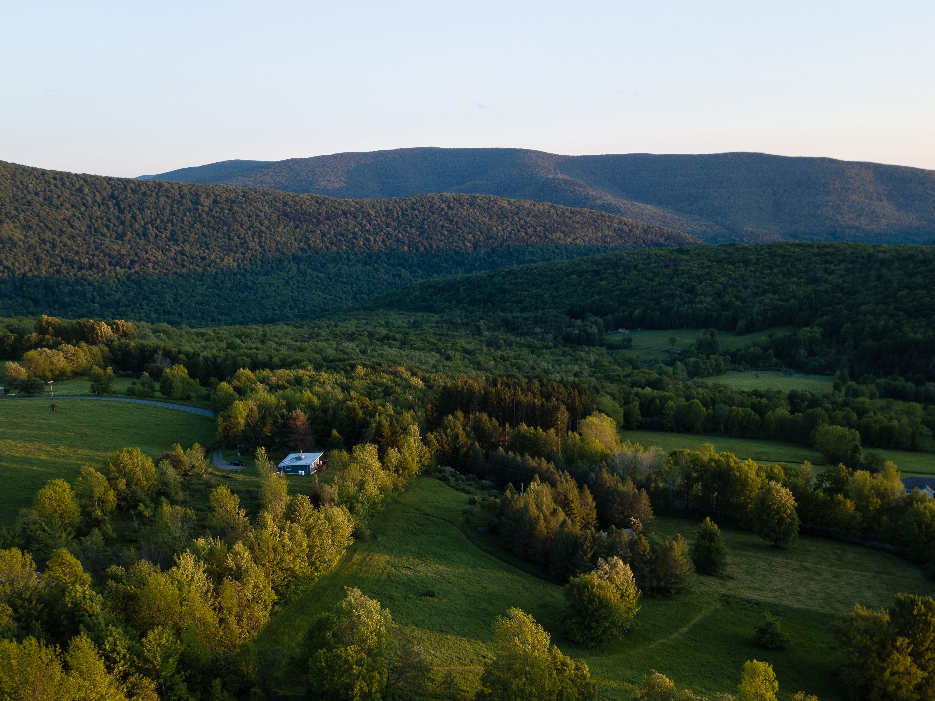 Paesaggio rurale con dolci colline, fitti boschi e una piccola casa con vialetto.