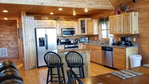 A cozy cabin-style kitchen with wooden cabinets, a refrigerator, microwave, stove, and a dishwasher. There are two bar stools and a wooden table with a woven placemat. - Long Range Views! 3 Fireplaces-Sleeps 20-Blue Ridge-Pool Table/HotTub-Pets OK. (Blue Ridge)