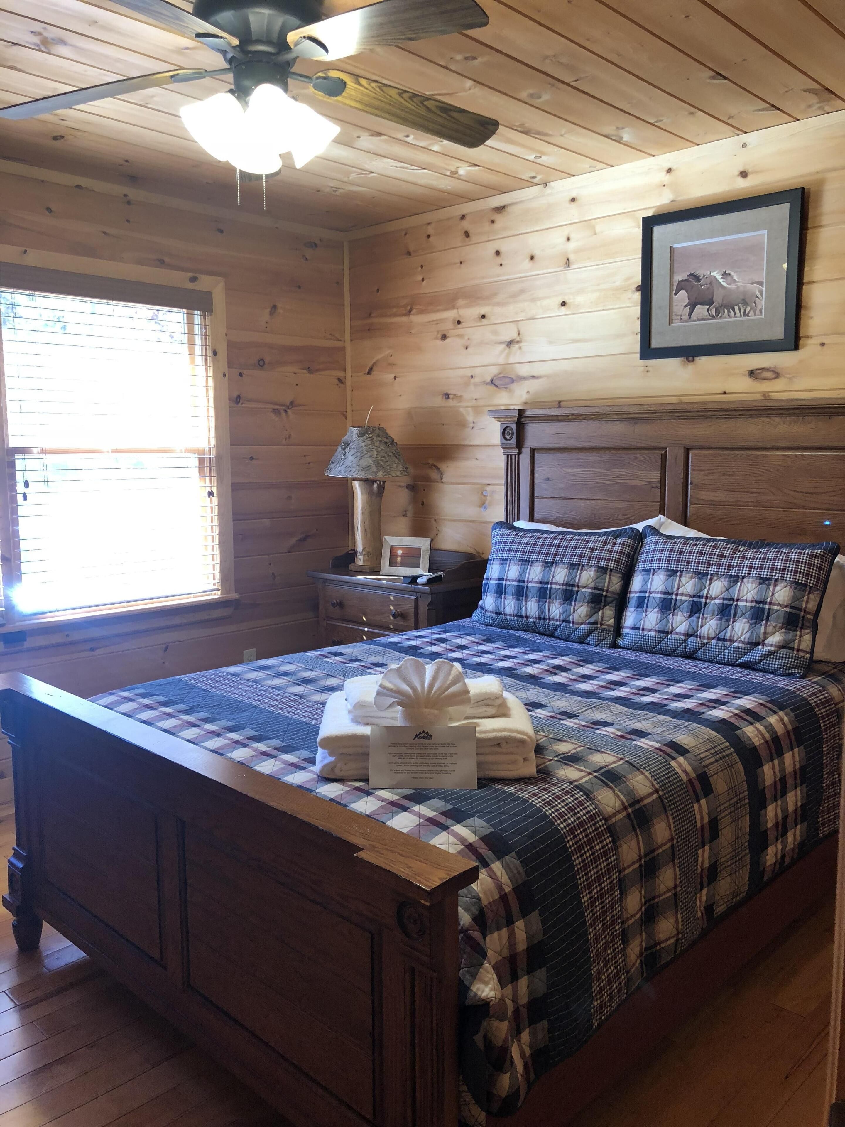 A wooden cabin-style bedroom with a bed, a ceiling fan, a window with blinds, a nightstand, and a framed picture on the wall.